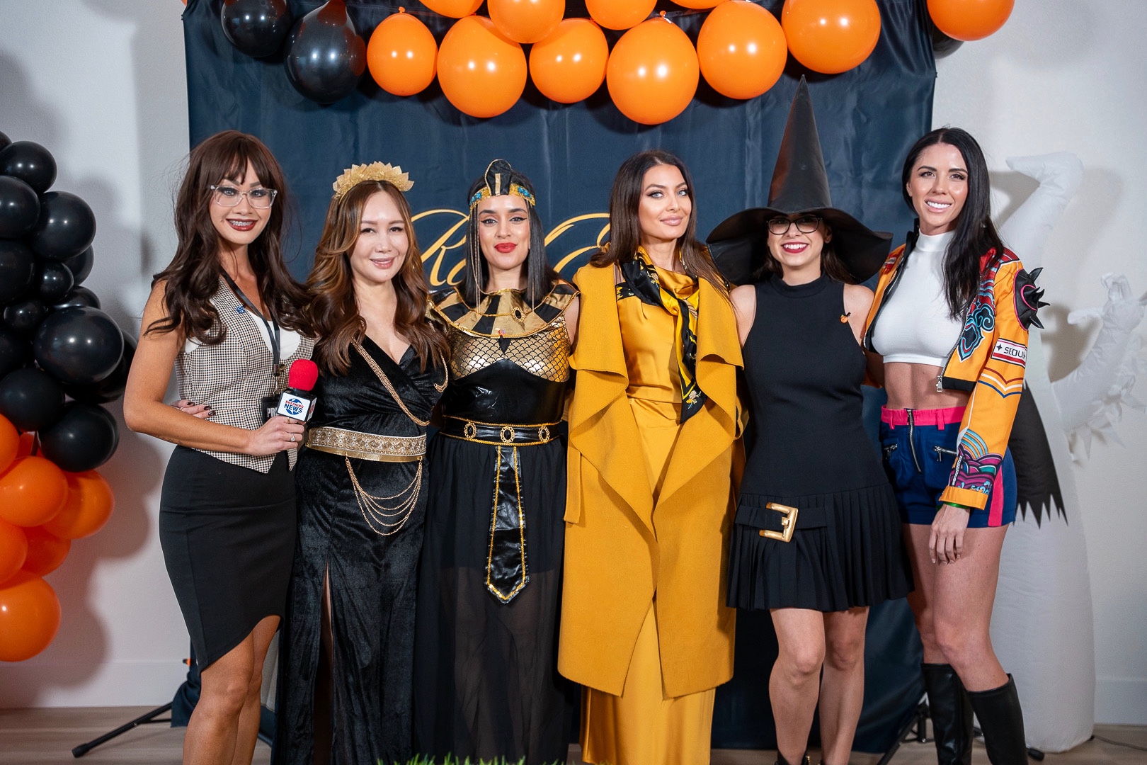 Six costumed friends posing at the REFC backdrop, from a reporter holding a BREAKING NEWS microphone to a witch in a pointed hat, beneath orange and black balloons