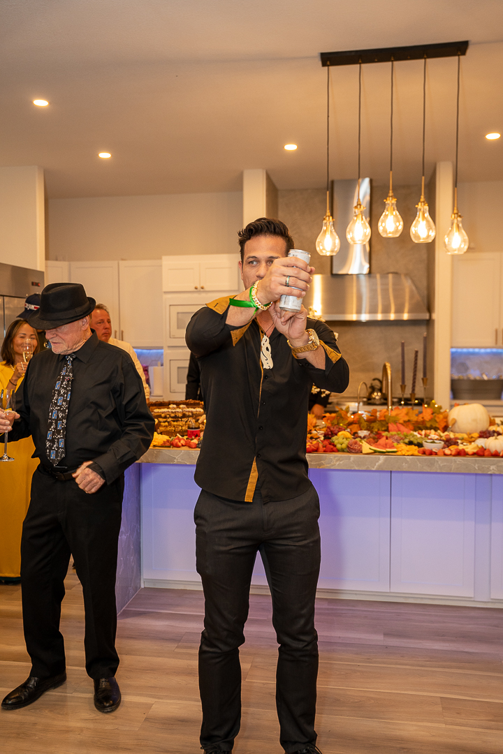 A guest in a black hat raising his drink in the kitchen