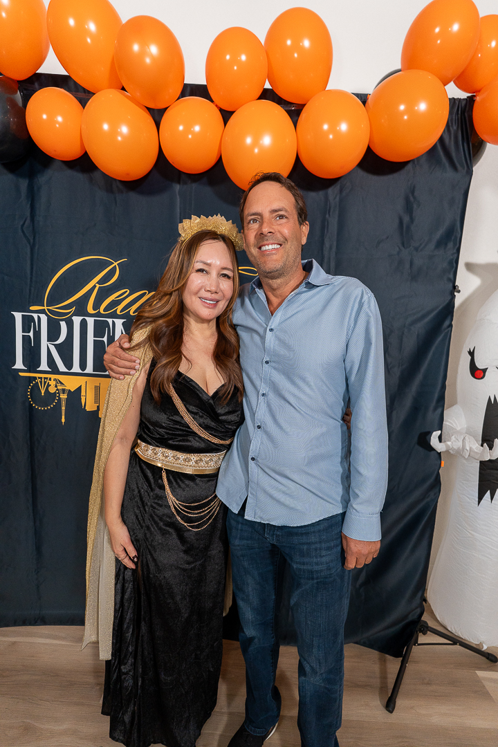 Two smiling friends posing at the REFC backdrop beneath the orange and black balloons