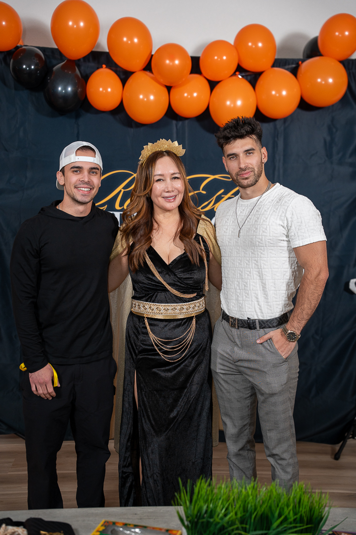 Three smiling guests posing beneath the orange and black balloons at the REFC backdrop